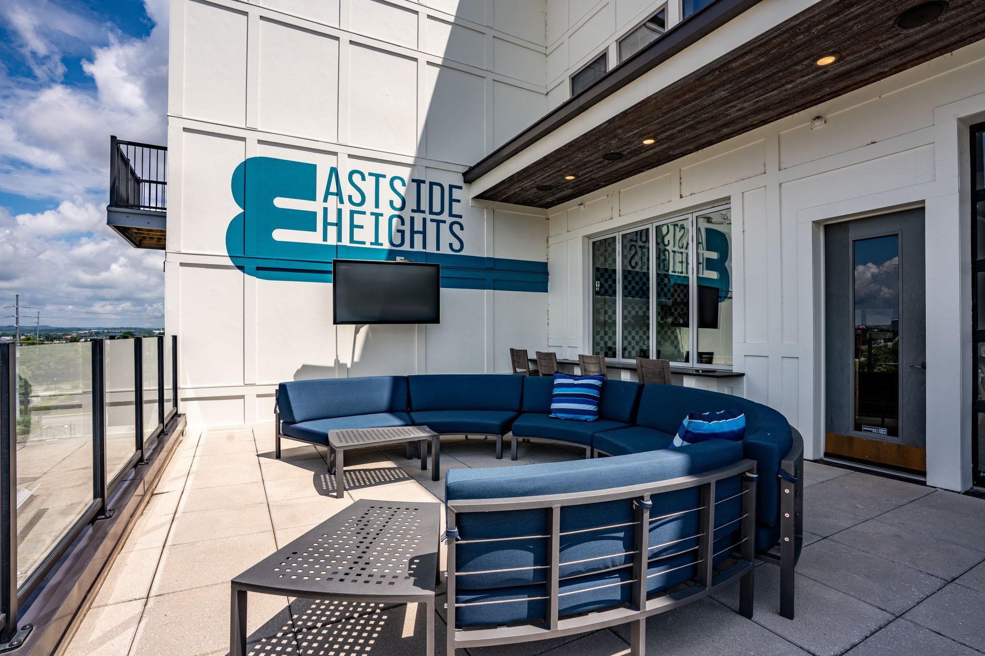 Outdoor lounge area with blue sectional sofa, coffee tables, TV, and signage for 