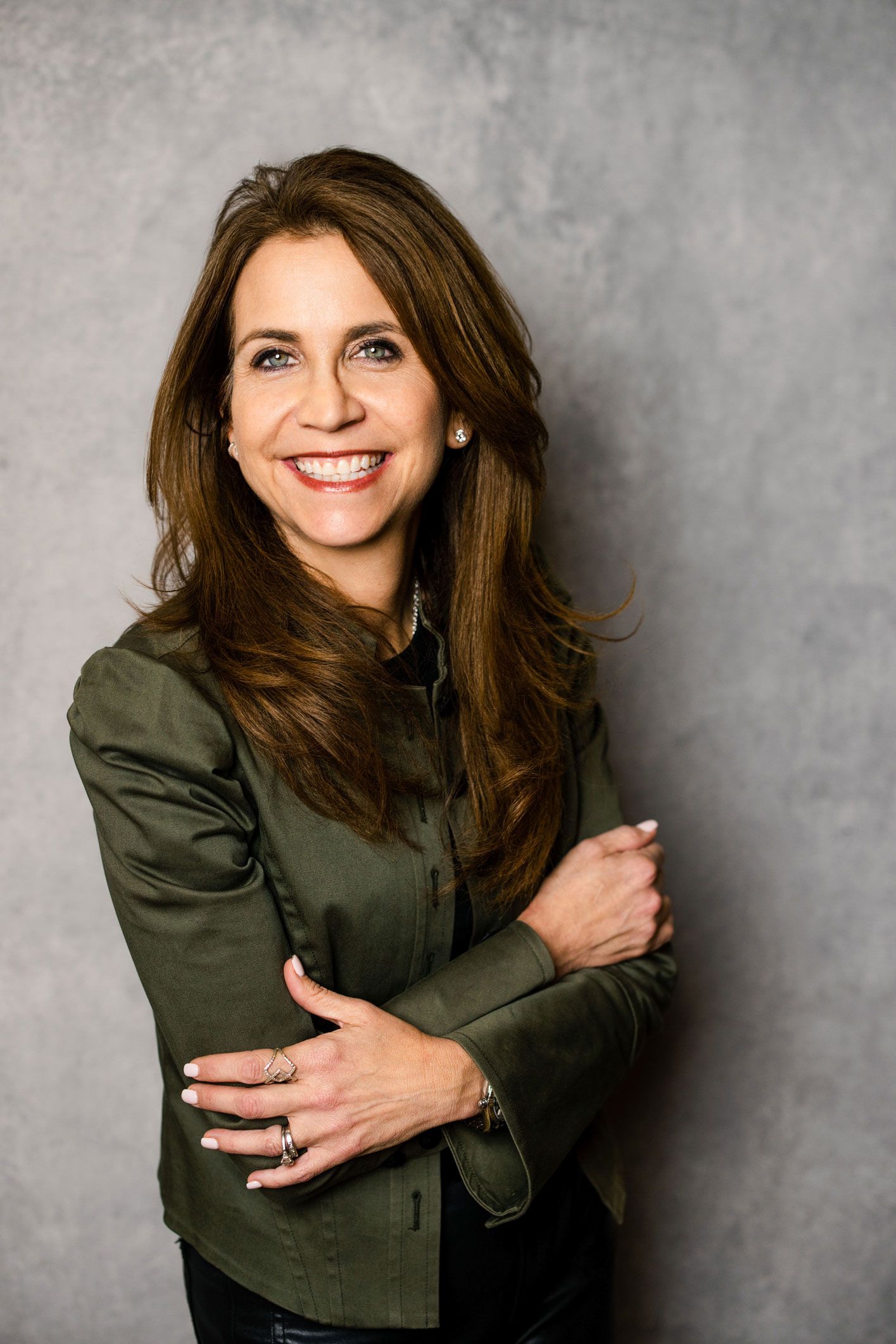 A smiling woman with long brown hair, wearing a dark olive jacket, standing against a grey textured background.