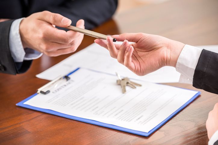 Two people in business attire exchange a pen over a contract and keys on a wooden table.