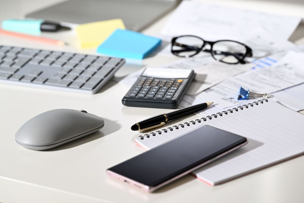 A desk with a cell phone , calculator , mouse , notebook , pen and glasses.