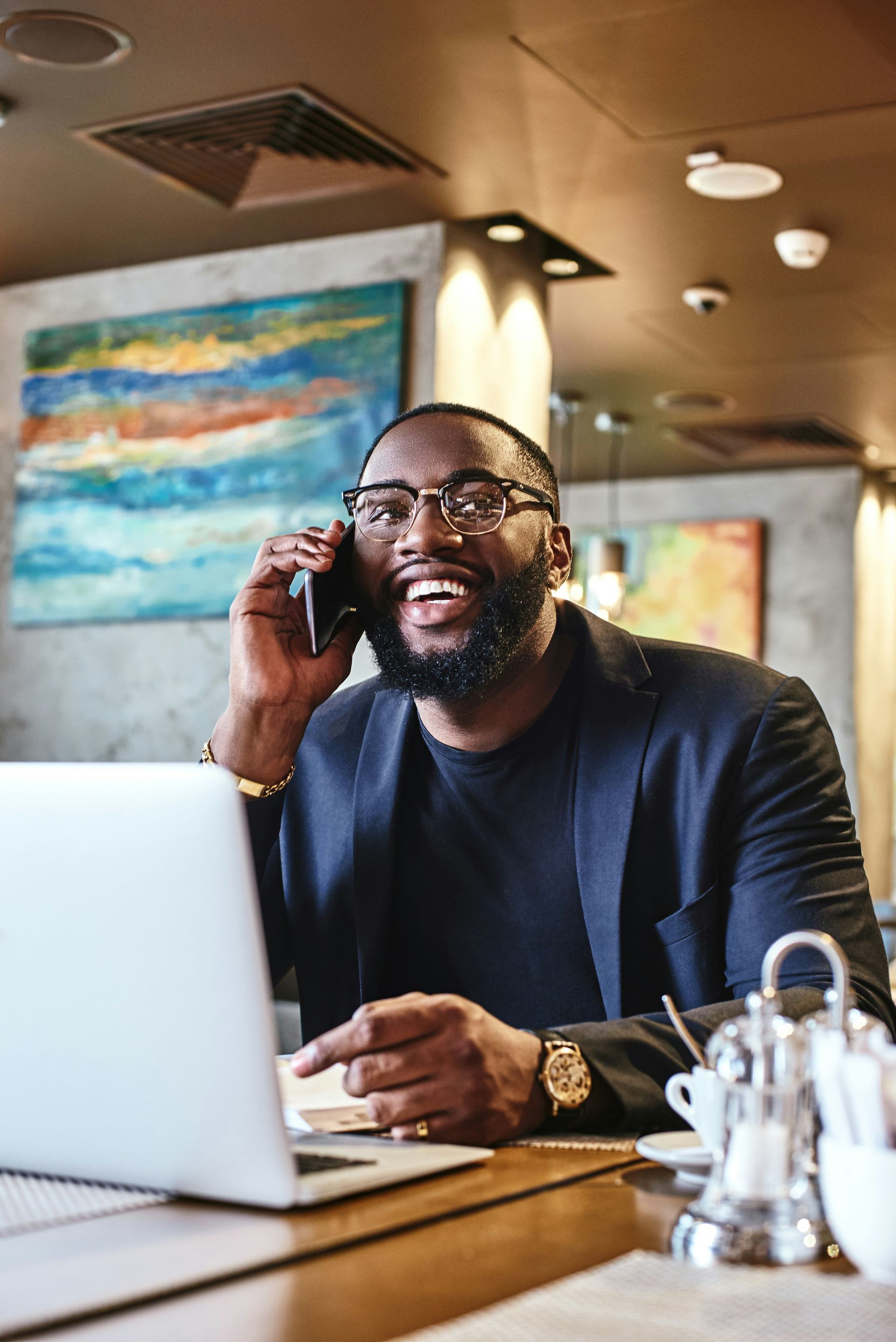 A man is sitting at a table with a laptop and talking on a cell phone.