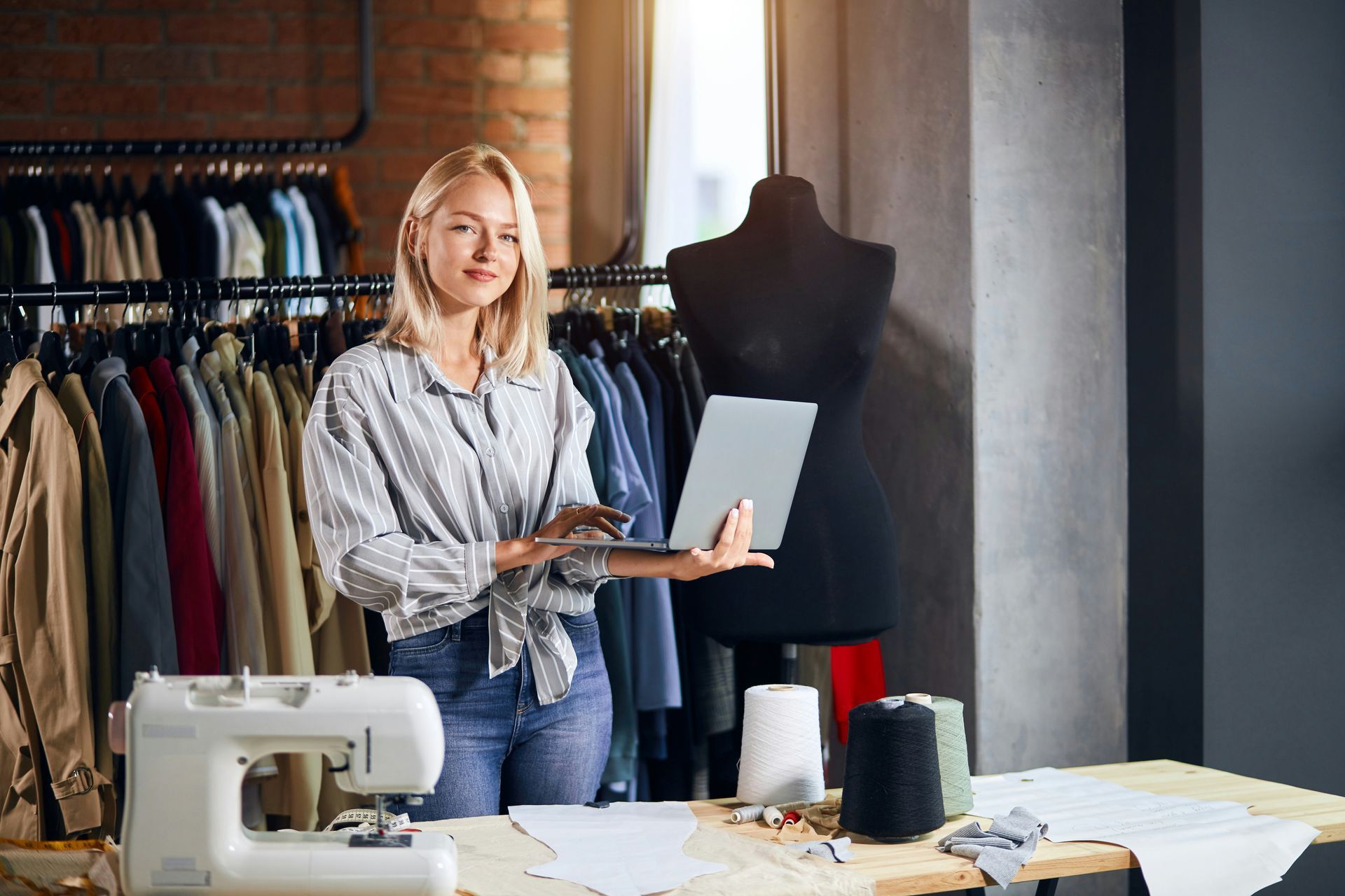 A woman is standing in front of a sewing machine holding a laptop.