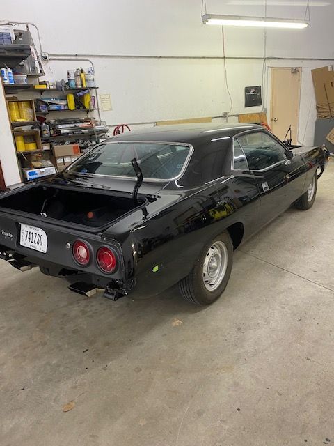 Black classic car in garage, trunk open, license plate visible, tools and shelves in background.