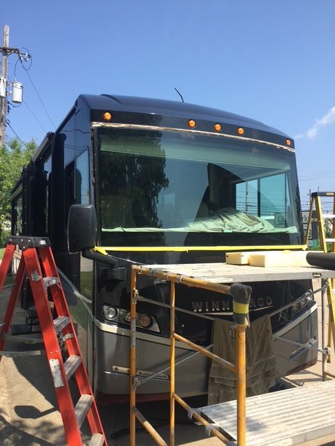 Black RV with taped windshield, on a sunny day, surrounded by scaffolding and a ladder.