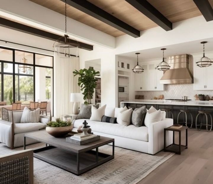 Open-concept living space with white sofas, dark beams, and a kitchen with white cabinets and metallic hood.