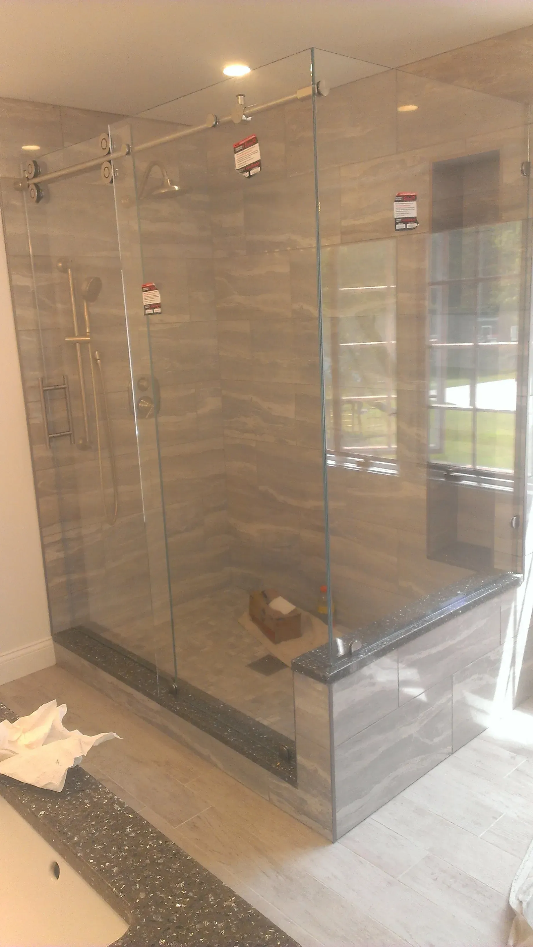 Glass-enclosed shower with gray tiled walls and floor, black trim, and a window in the background.