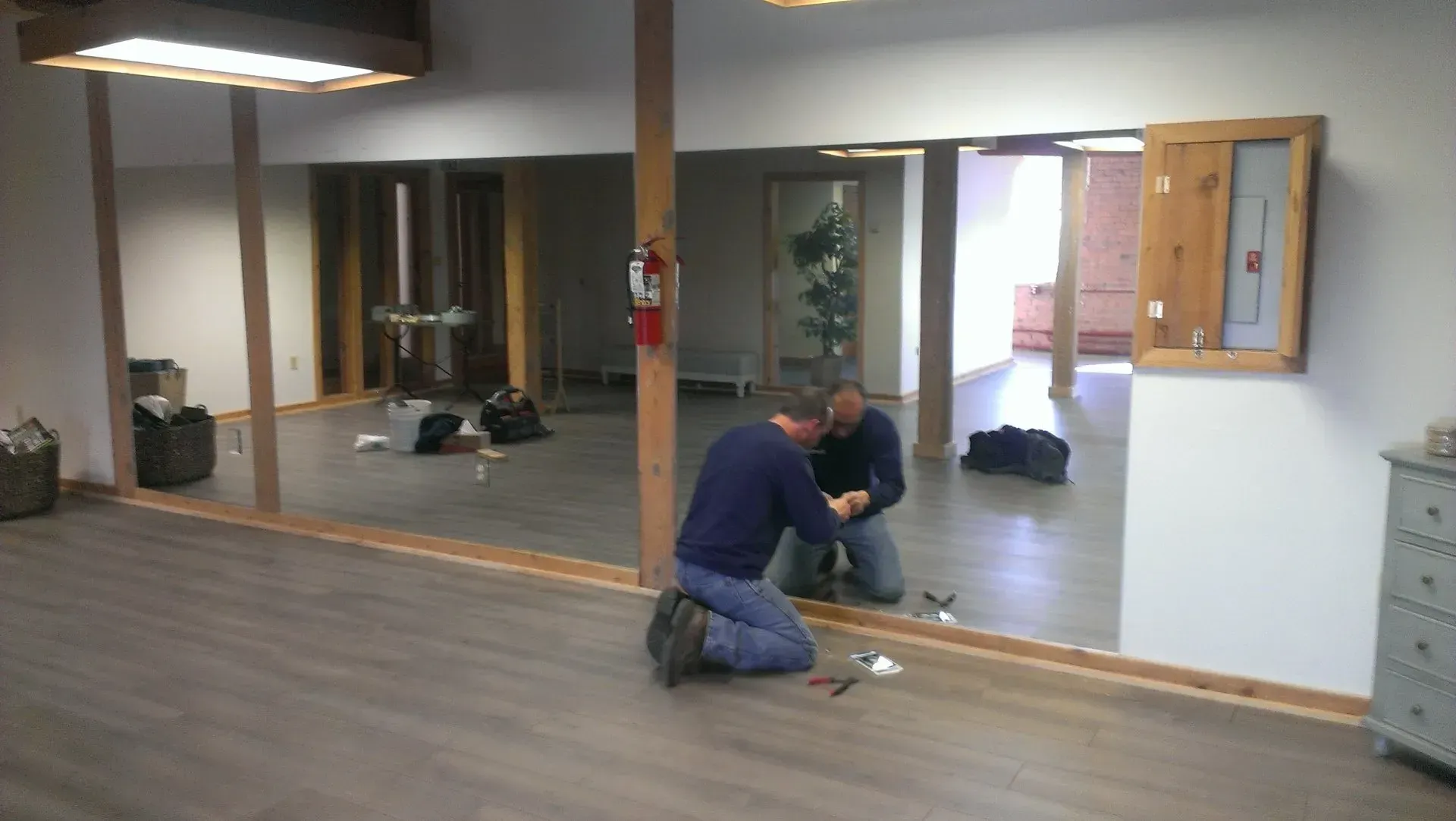 Two men installing a mirror on a wall in a room with wood floors and unfinished doorways.