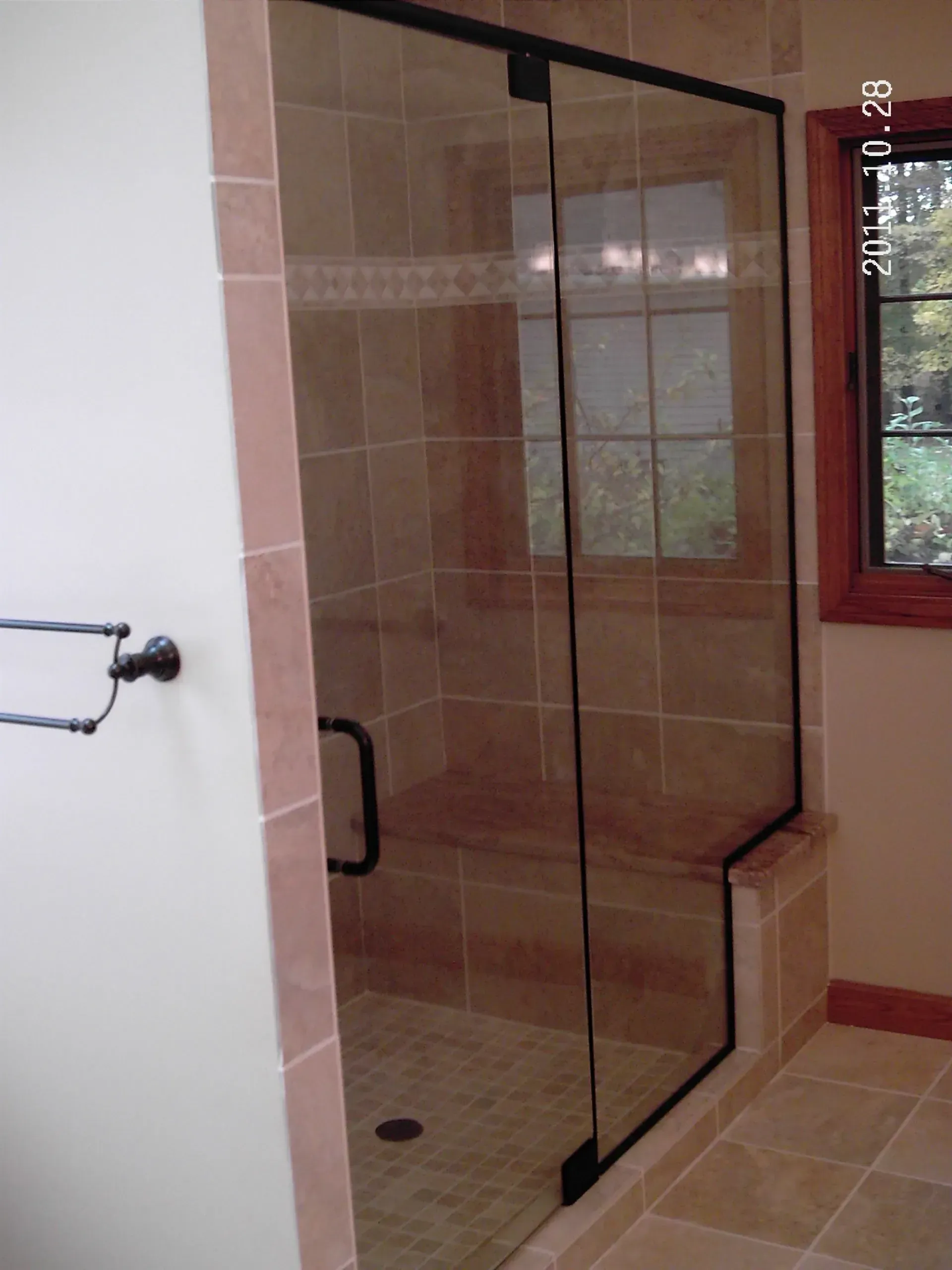 Shower with glass door, tan tile, and a bench. Black framed door and handle. Window in background.
