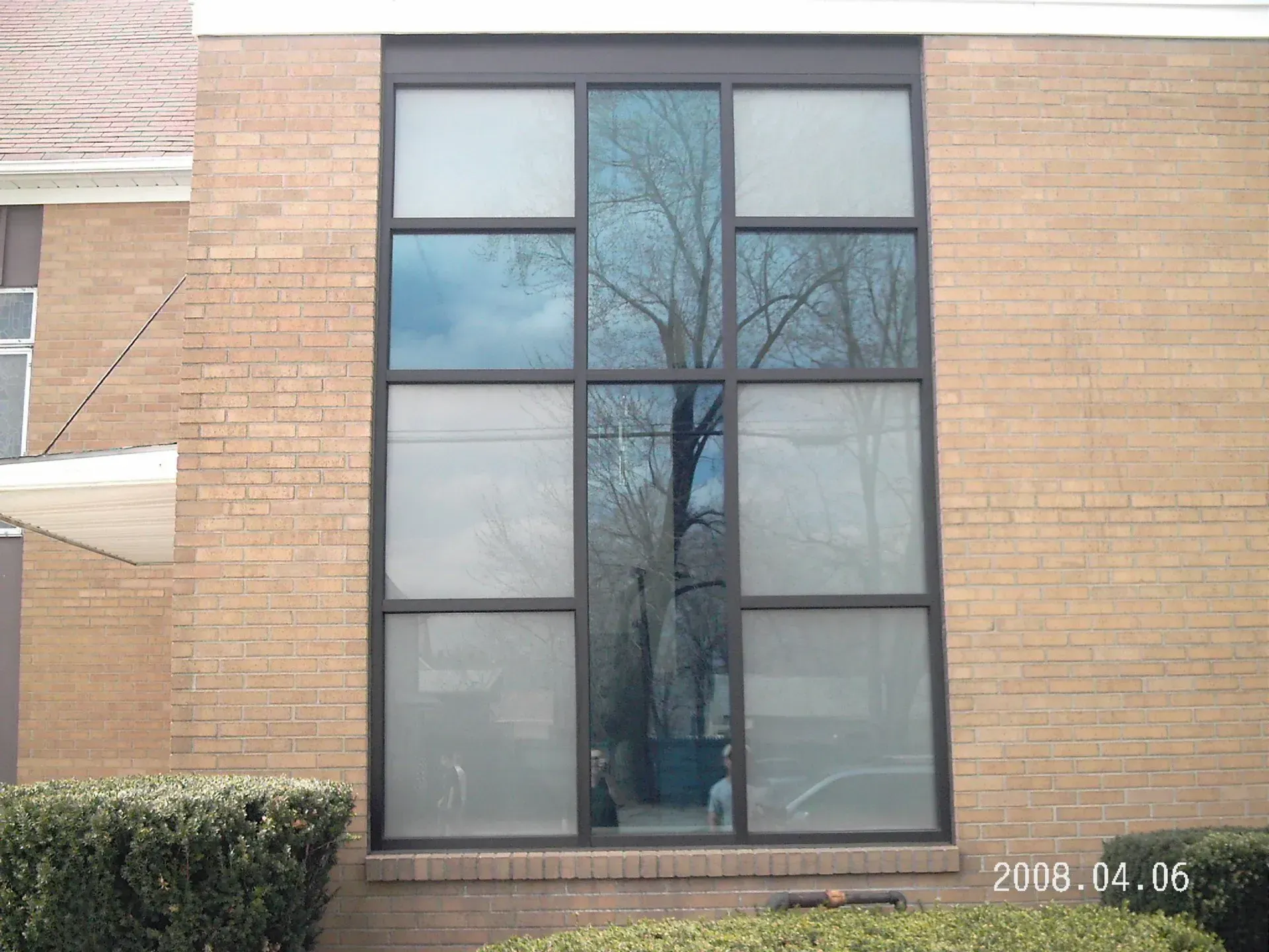 Large window of a brick building reflecting a tree, sky, and building's interior.