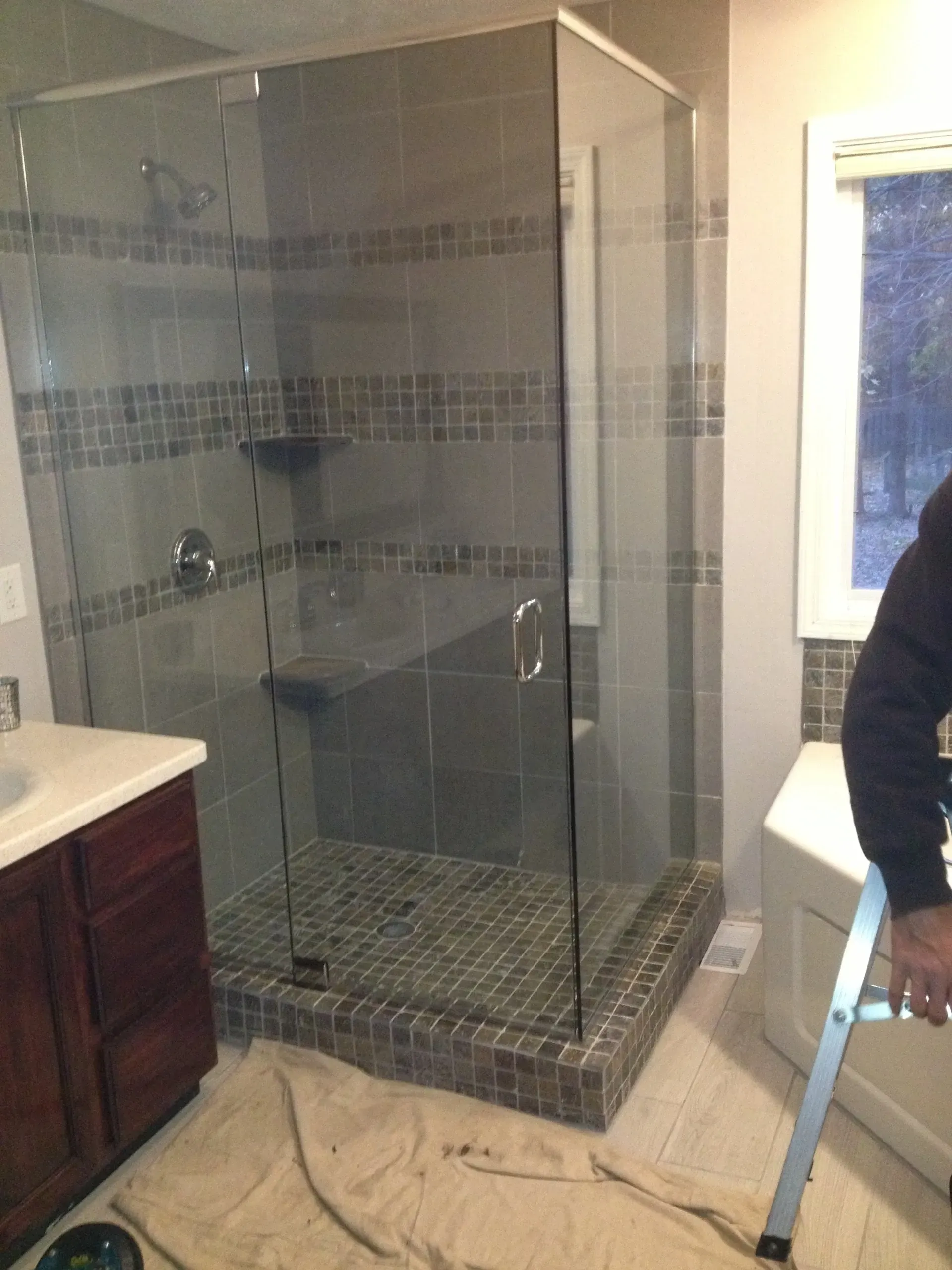 Glass shower enclosure with gray tile, installed in a bathroom. A person stands nearby holding a ladder.