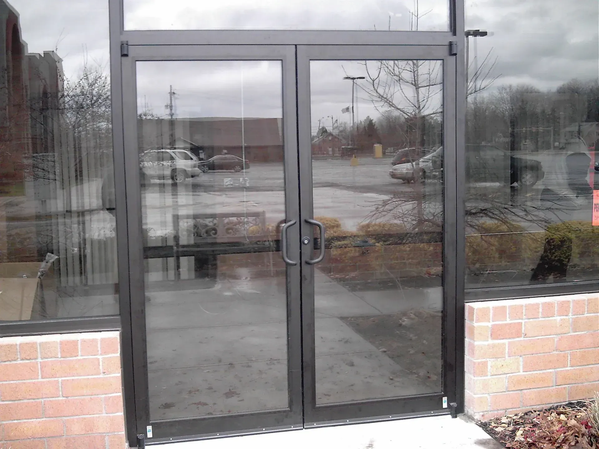 Double glass doors with black frames. A parking lot and cloudy sky are reflected.