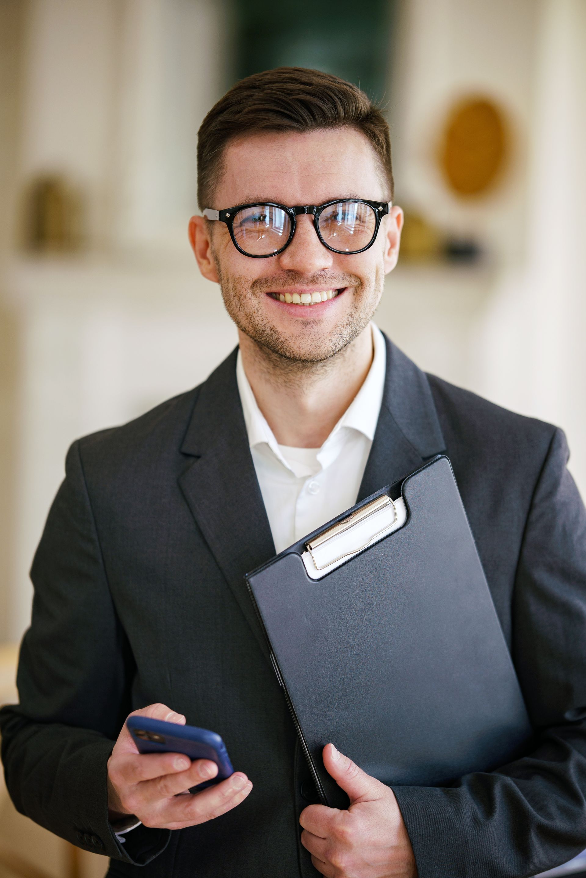 Man in suit, glasses, smiles, holds clipboard and phone in a bright room.