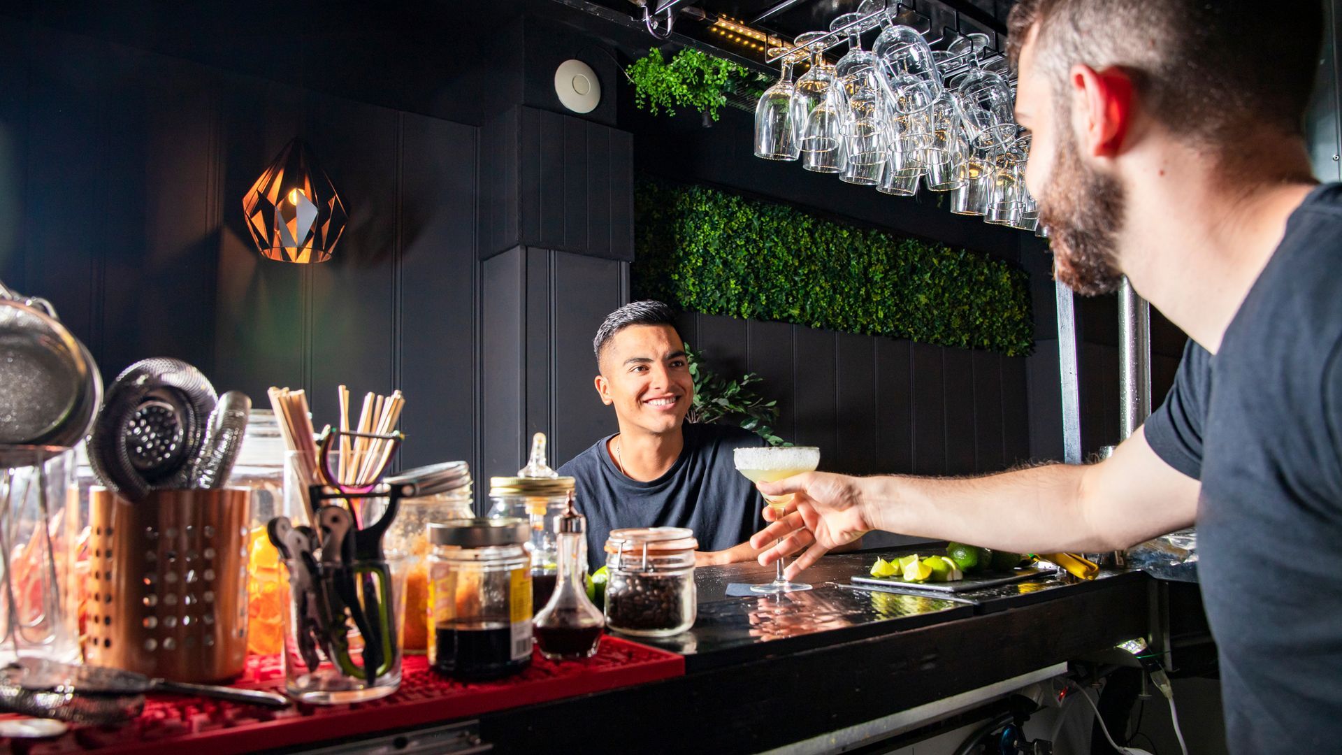 Bartender hands a cocktail to a smiling man at a bar. Dark interior, bar tools, green wall.
