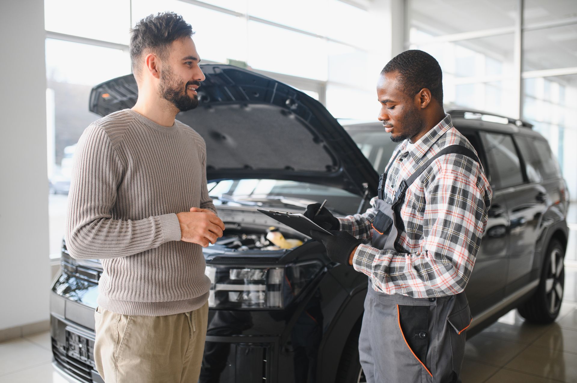 A car mechanic shows a customer a document, with a car and open hood in the background.