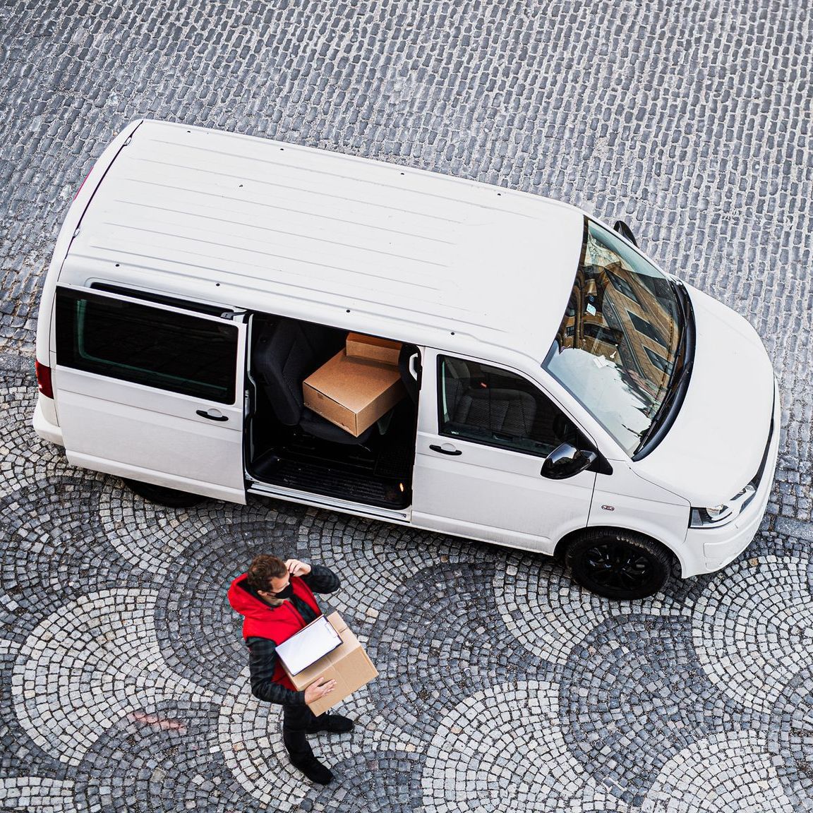 A person in red jacket carrying packages next to a white delivery van on cobblestone.