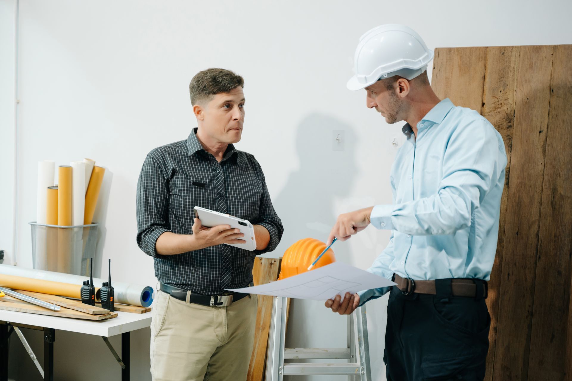 Two men in construction setting reviewing blueprints and tablet.