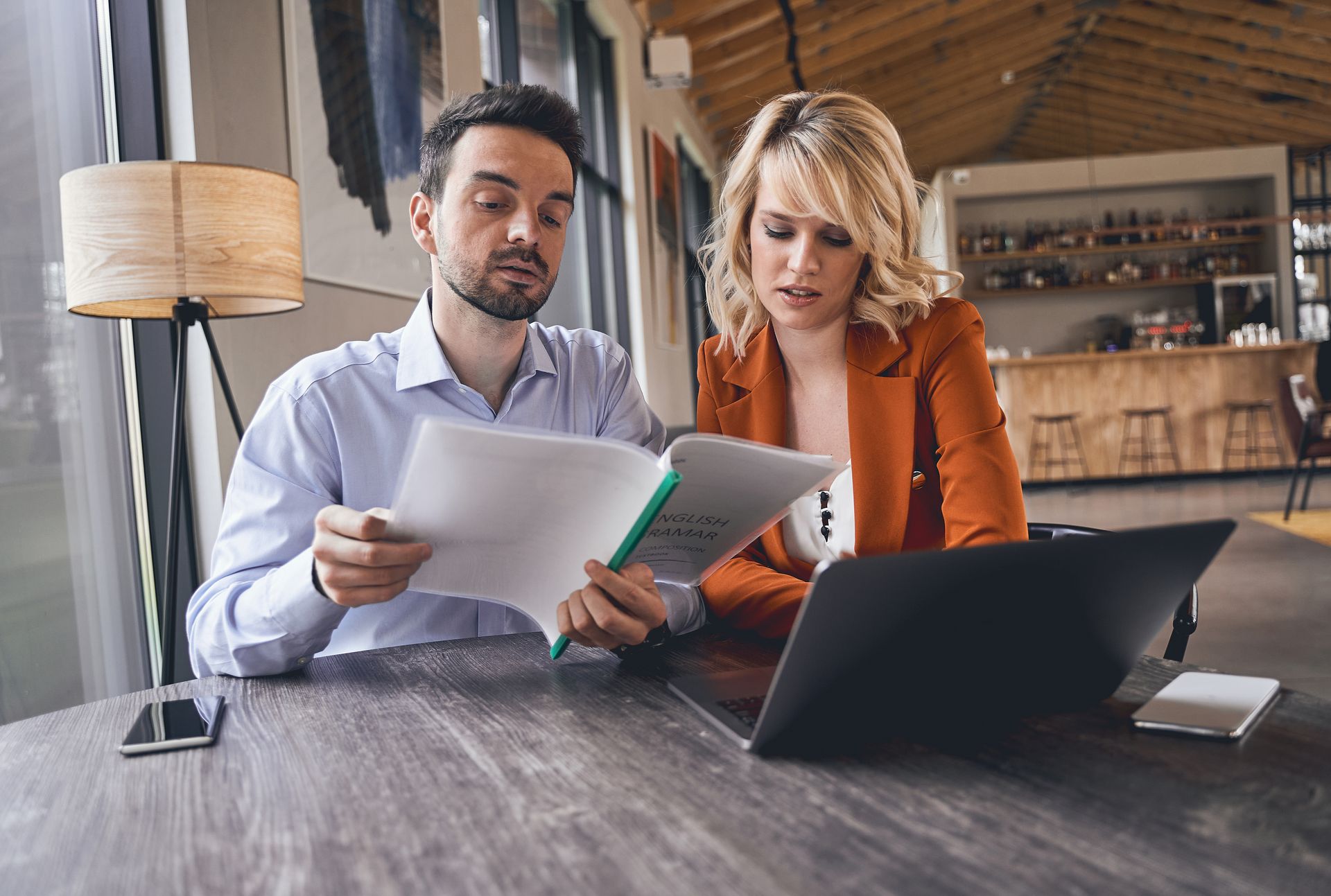 Man and woman reviewing documents together at a table with a laptop in a cafe setting.