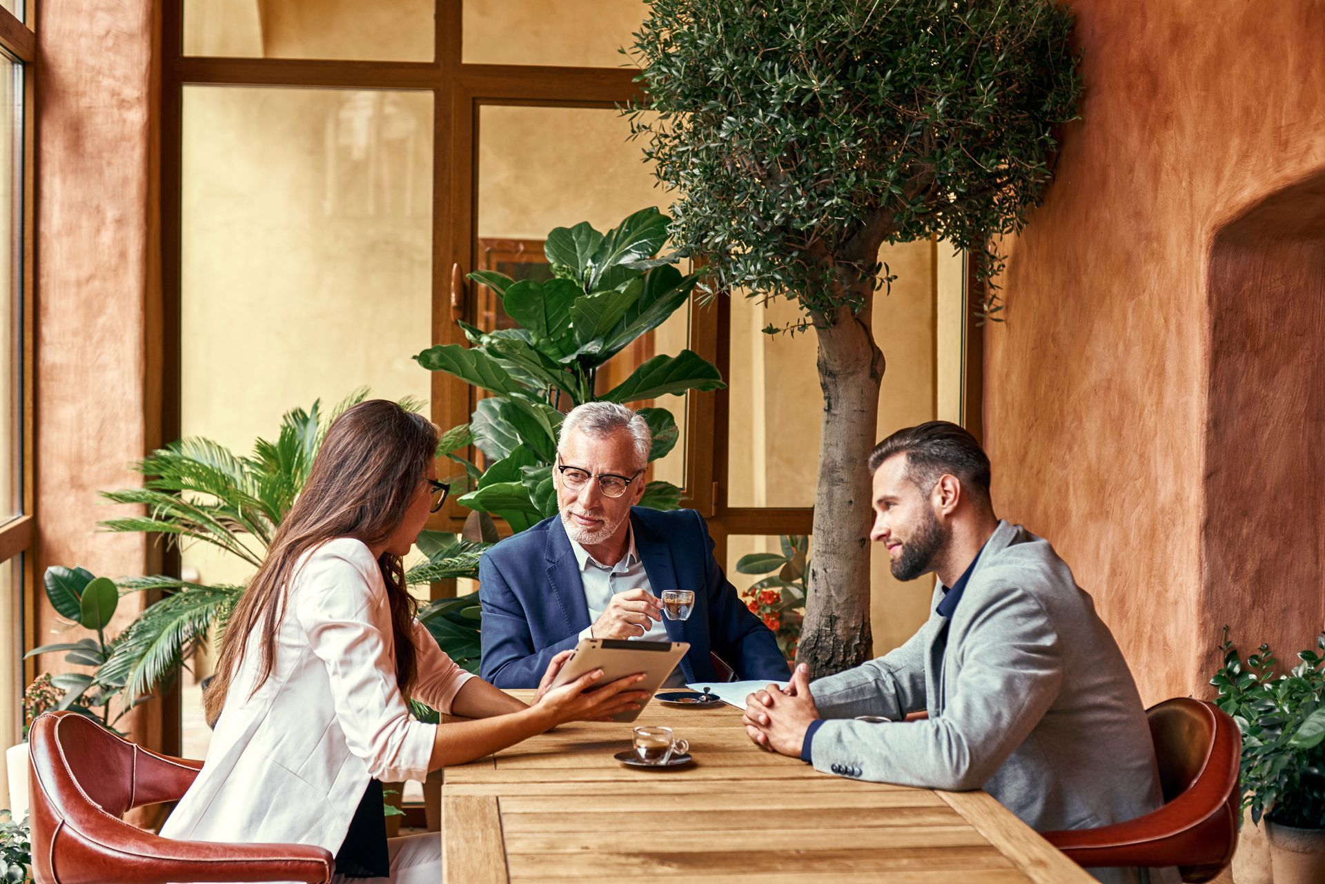 Three businesspeople at a wooden table, reviewing a tablet. Indoor, with plants.