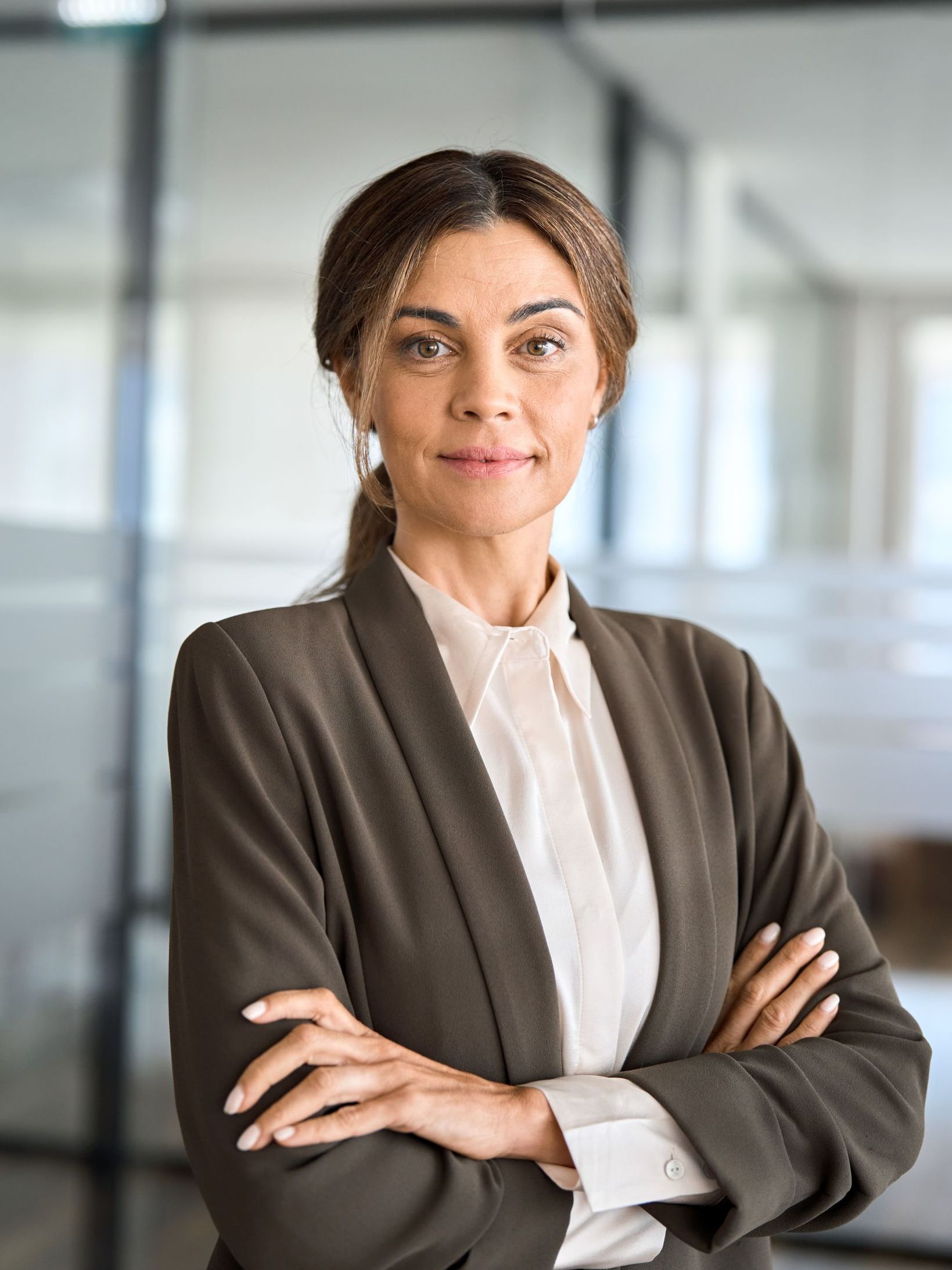 Woman in a business suit with arms crossed, smiling confidently, in a modern office.