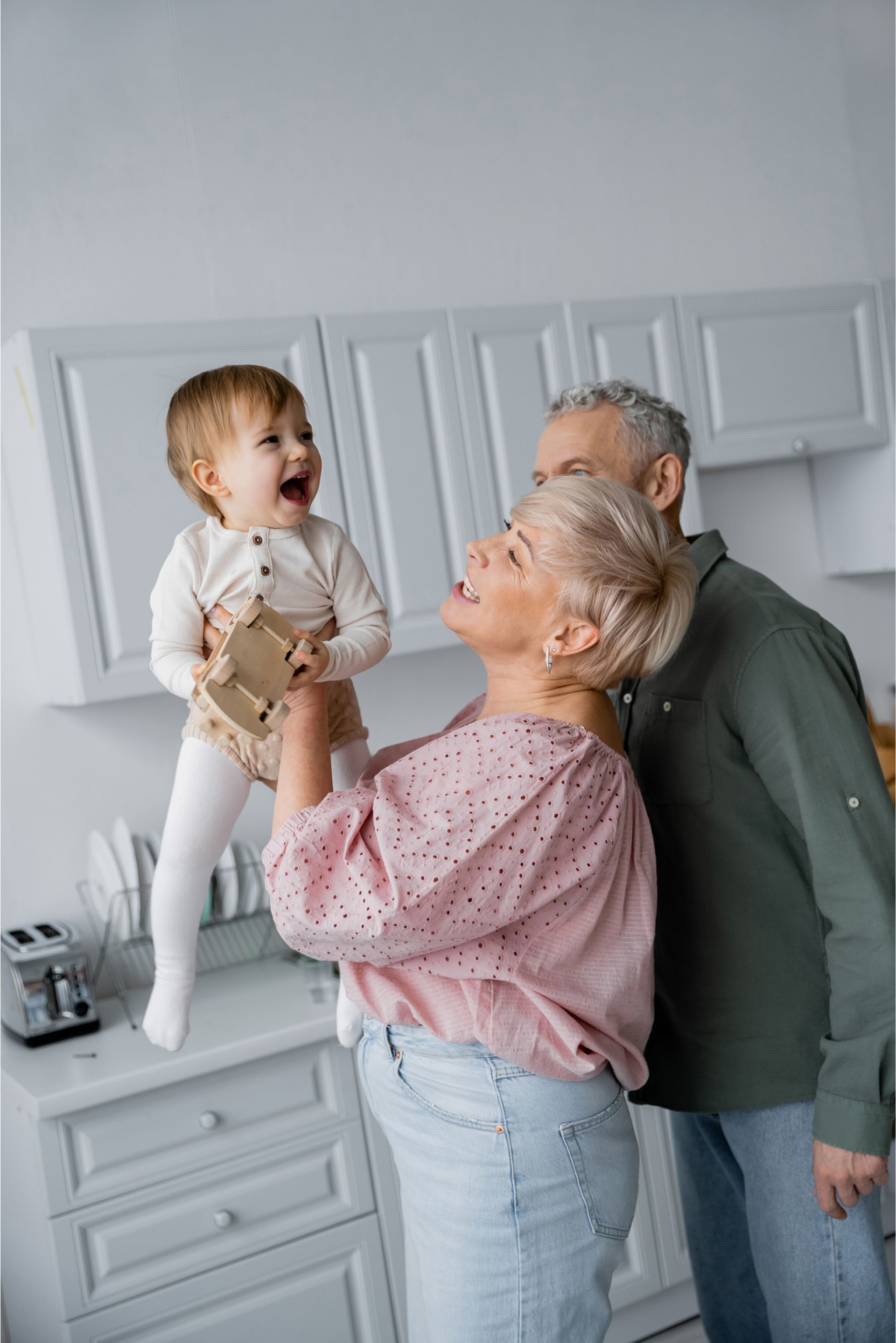 A laughing baby held by a smiling woman, with a gray-haired man looking on, in a kitchen.