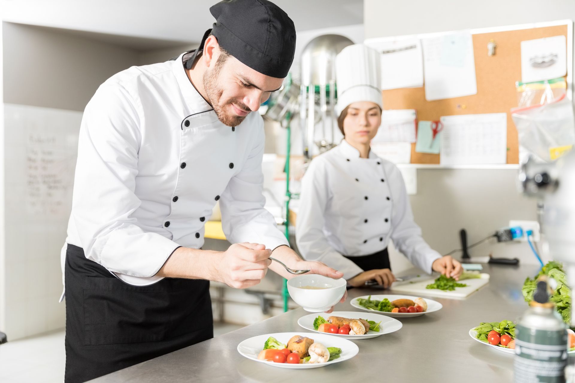 Two chefs plating food in a commercial kitchen; man sprinkles over plate; woman preps greens.