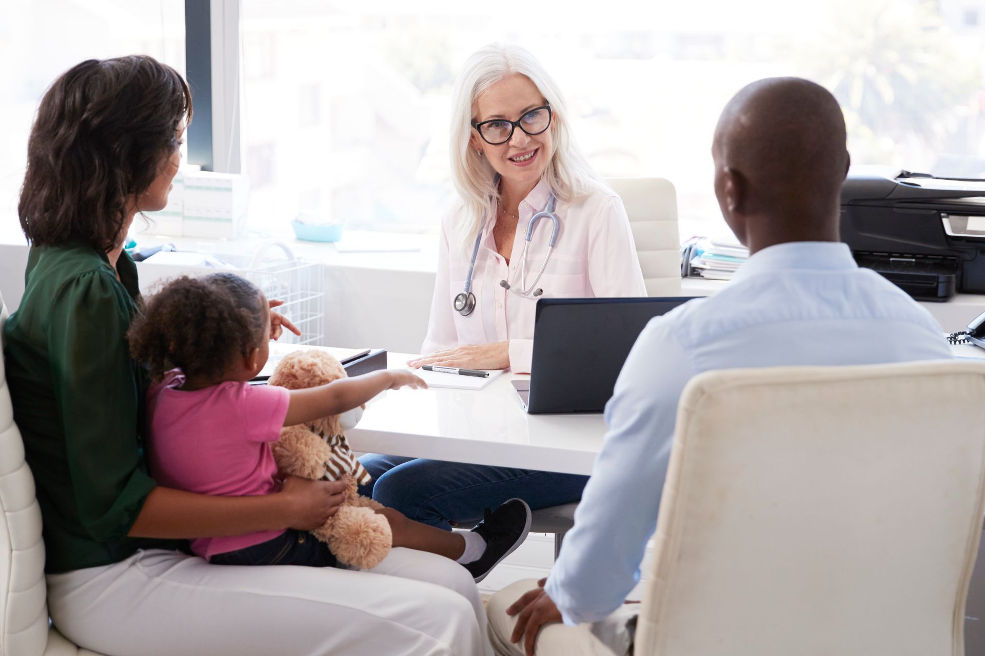 Family at doctor's office: parents with young child consult with a smiling, gray-haired doctor at a desk.