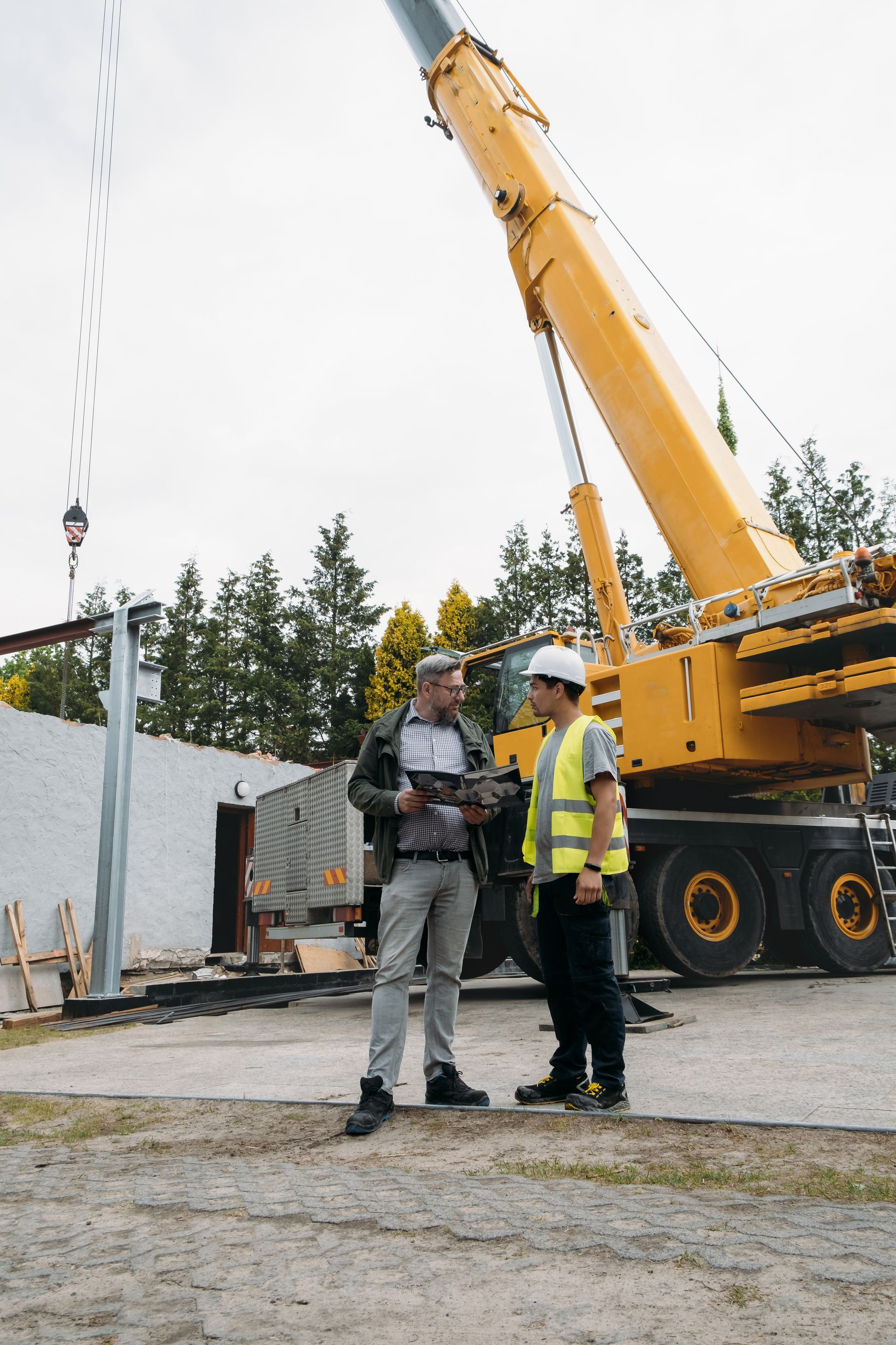 Two men at a construction site; a crane lifts metal beams. One man holds a tablet.