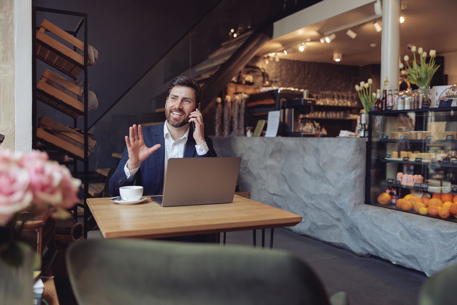 Man in suit at cafe, on phone, gesturing. Laptop and coffee on table.