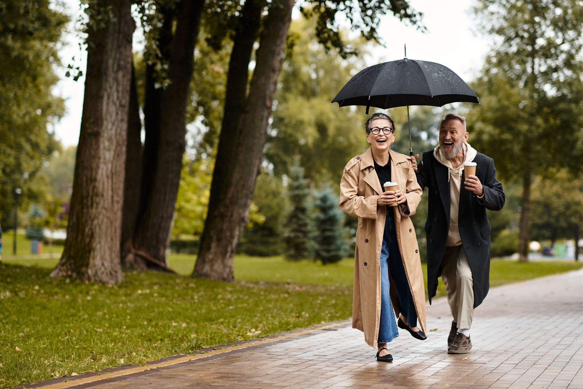 A couple walks in a park, sharing an umbrella, holding coffee, smiling.