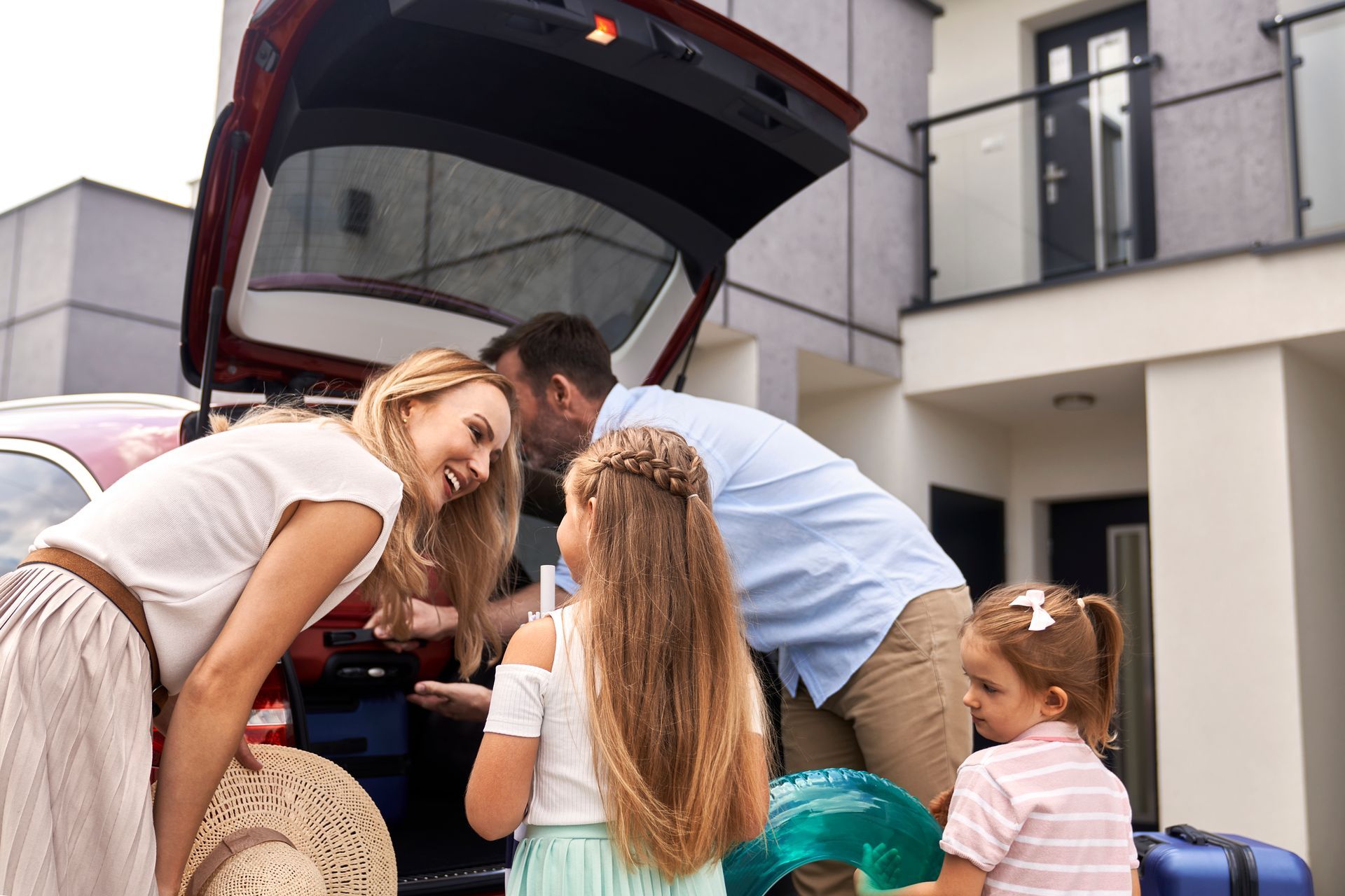 Family packing car trunk outside a house: mom, dad, two daughters.