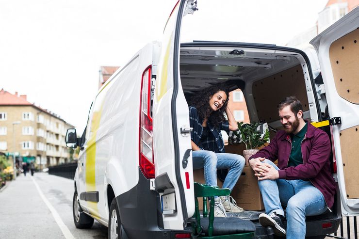 A smiling couple sits in an open van, boxes visible. Urban street setting.