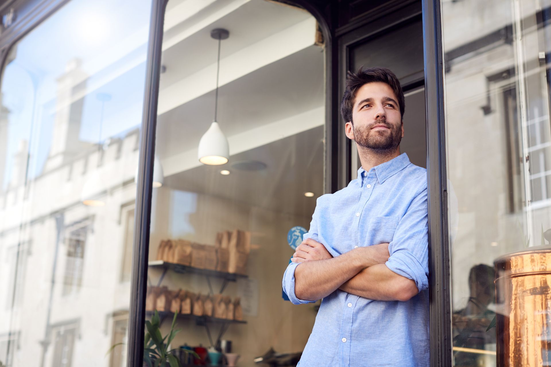 Man with crossed arms stands in shop doorway, looking up thoughtfully.