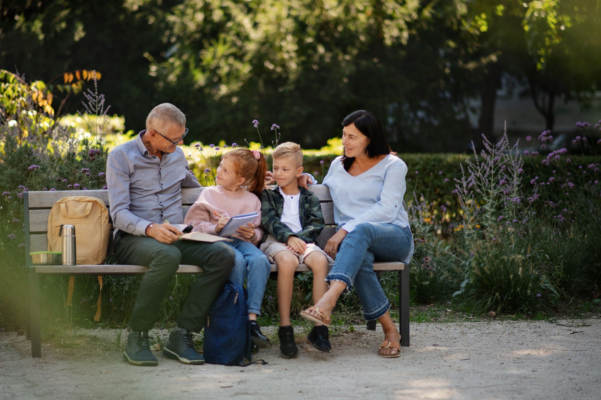 Family of four sits on a park bench, reading and relaxing together. Sunlight, outdoors.