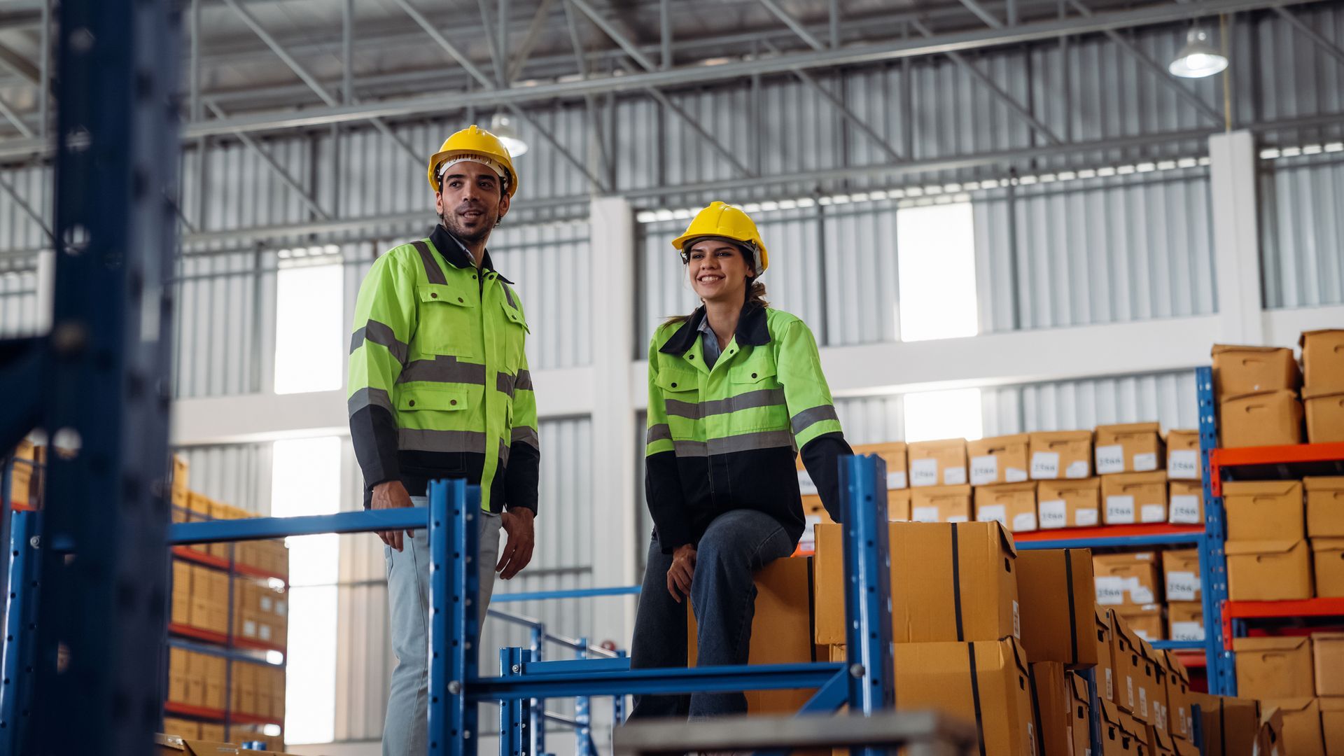 Two warehouse workers in safety vests and hard hats, standing near boxes in a large warehouse.