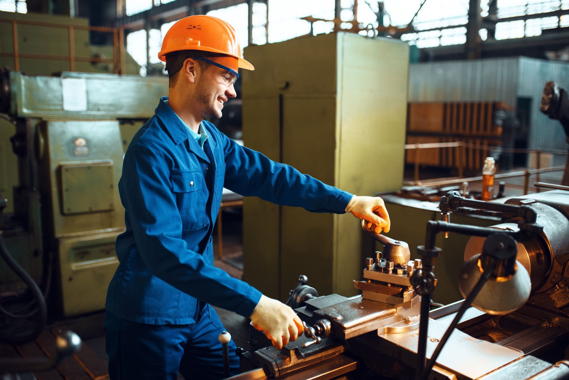 Man in blue overalls and orange hard hat operating a metal lathe in a factory.