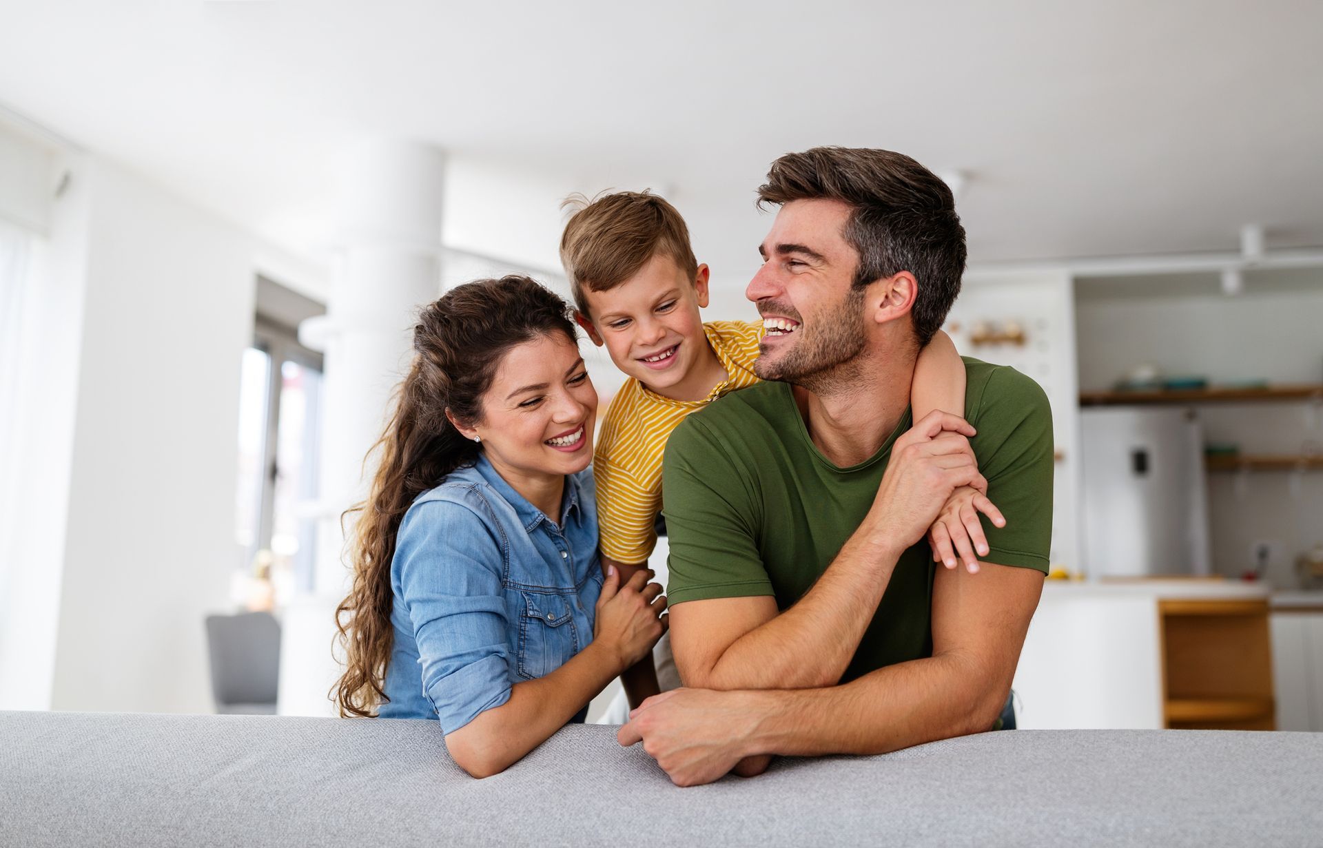Family of three smiling, embracing each other inside a home, with the child hugging the father.