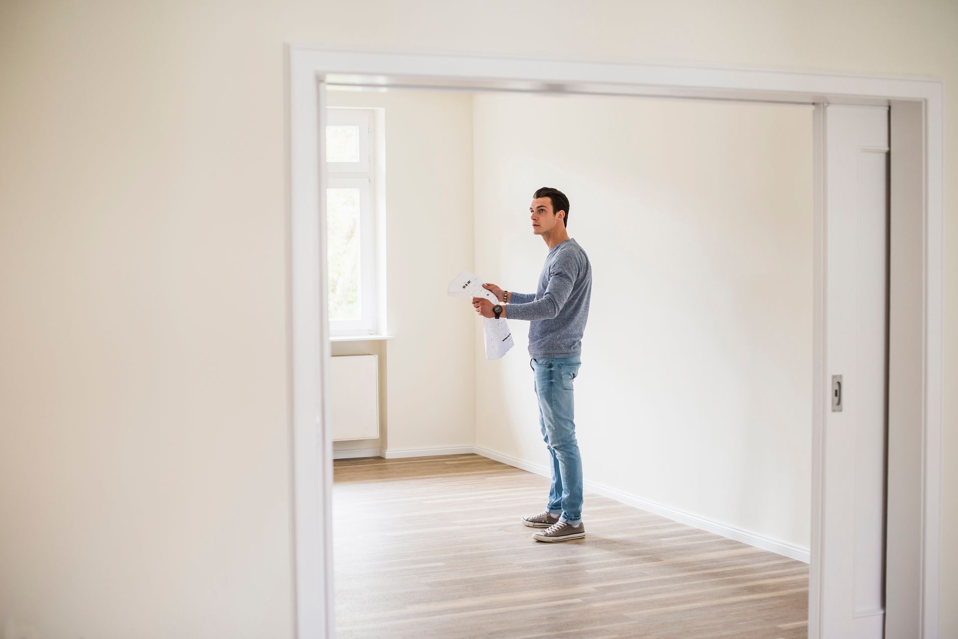 Man in blue jeans and sweater stands in a bright, empty room, holding papers.