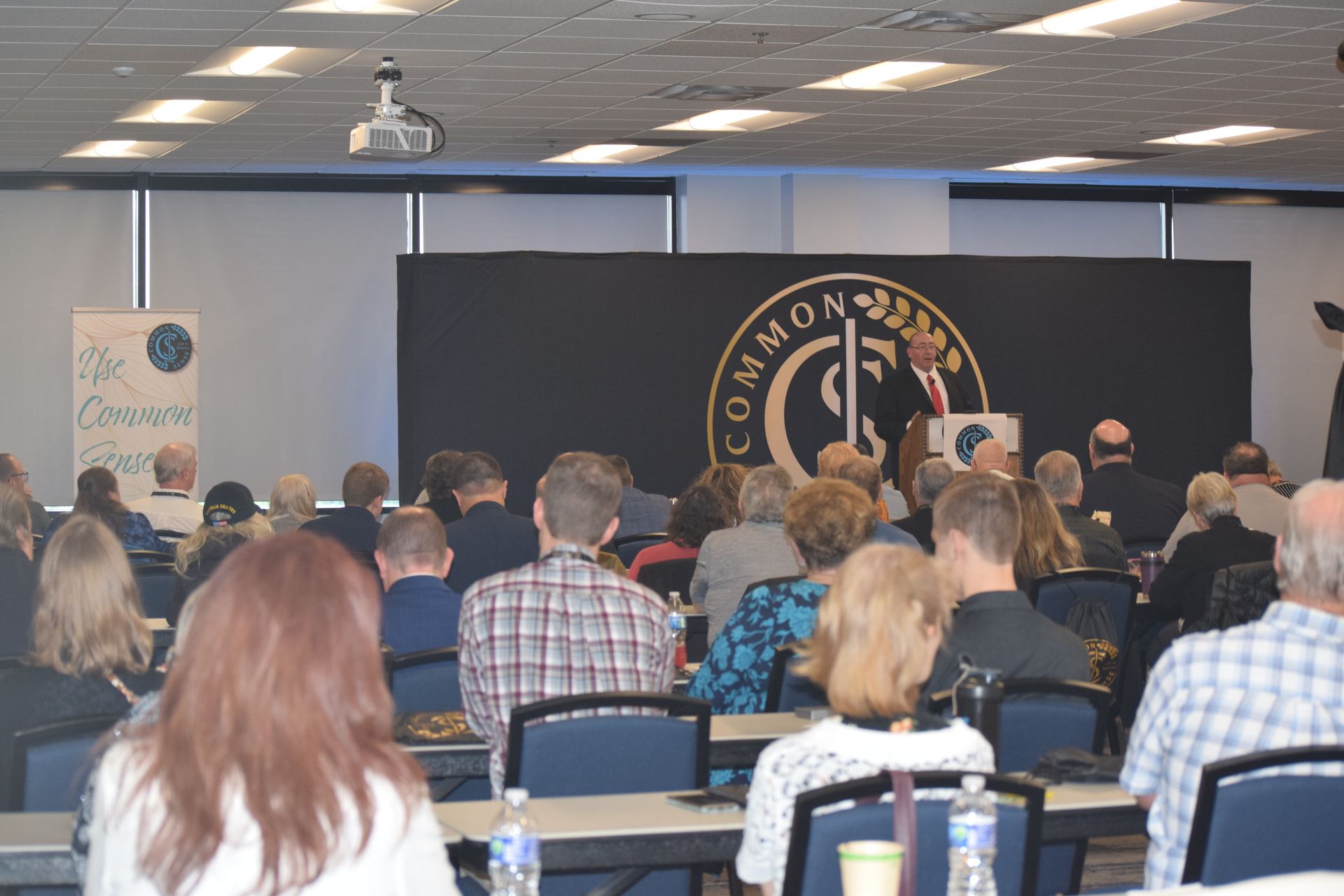 A group of people are sitting in a room with a sign that says the commons