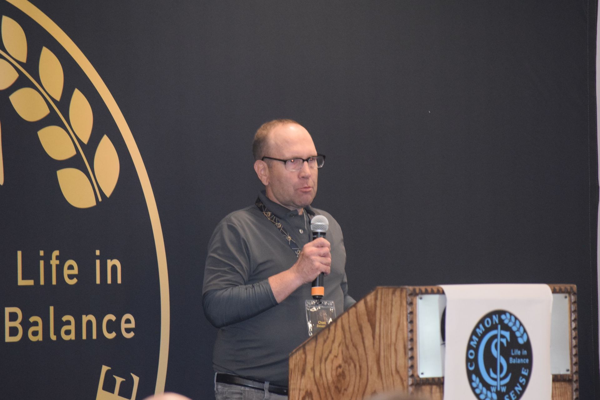 A man stands at a podium with a microphone in front of a sign that says life in balance