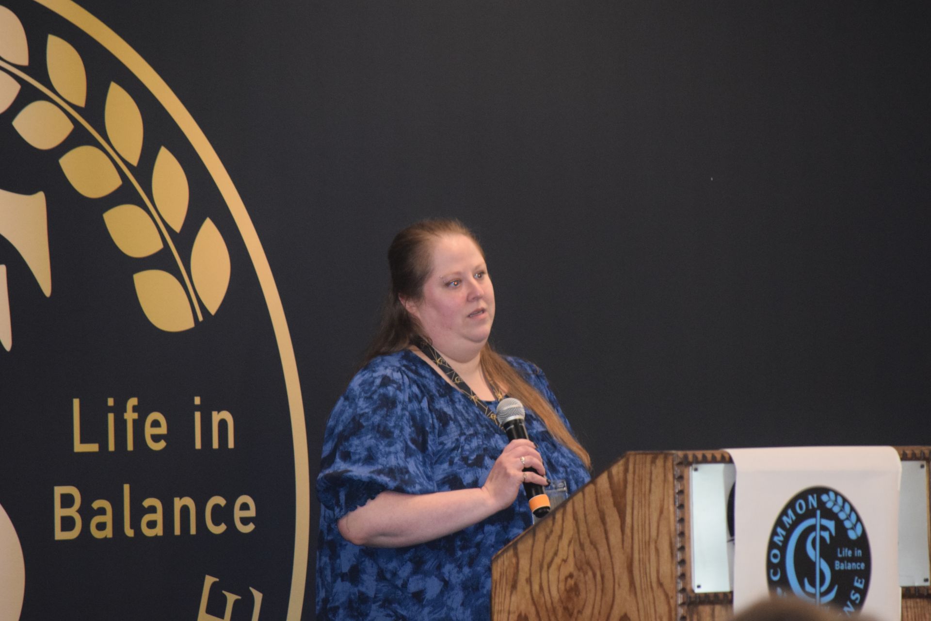A woman stands at a podium with a microphone in front of a sign that says life in balance