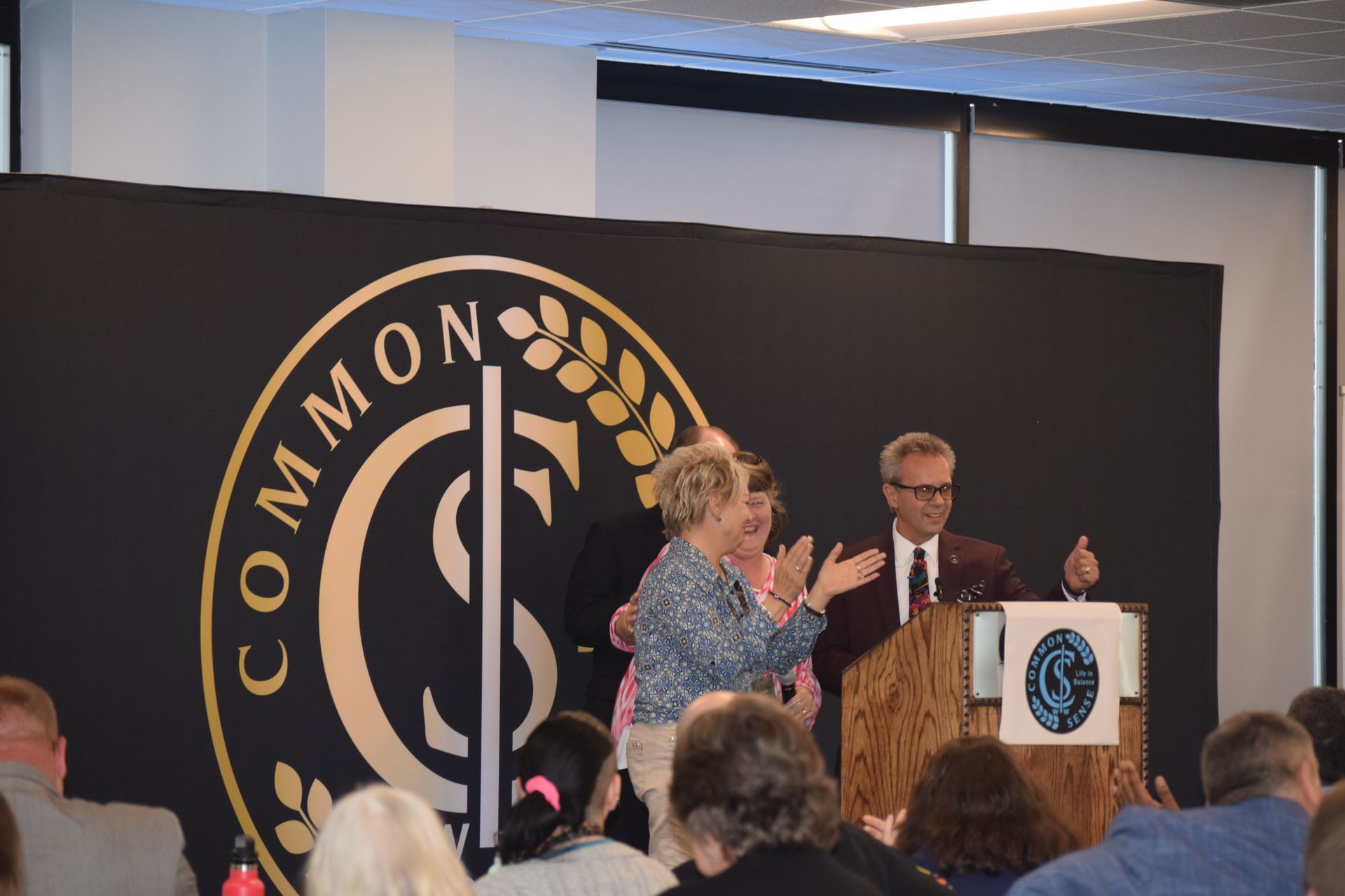 A man stands at a podium in front of a crowd with a sign that says common