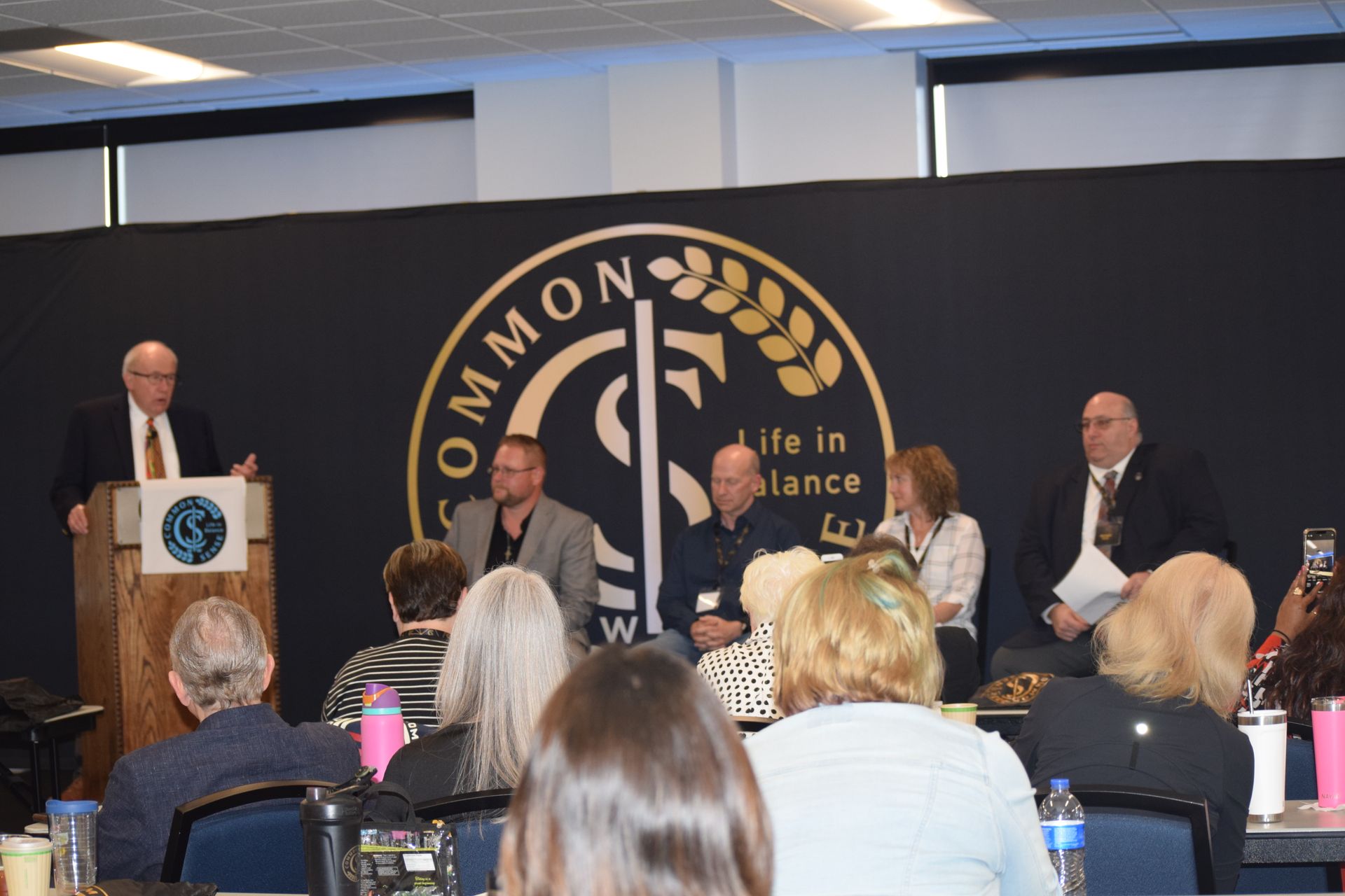 A group of people are sitting at tables in front of a sign that says common life in balance.