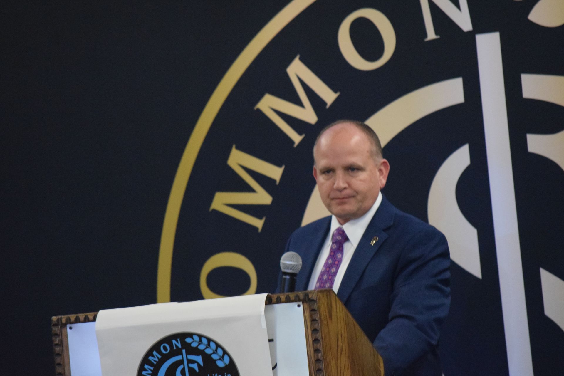 A man in a suit and tie stands at a podium in front of a sign that says common