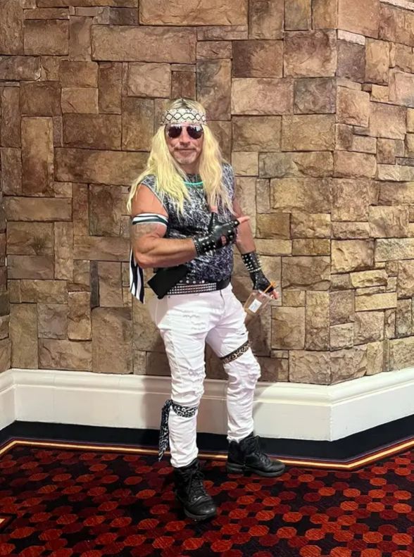 A man with blonde hair and sunglasses is standing in front of a brick wall.