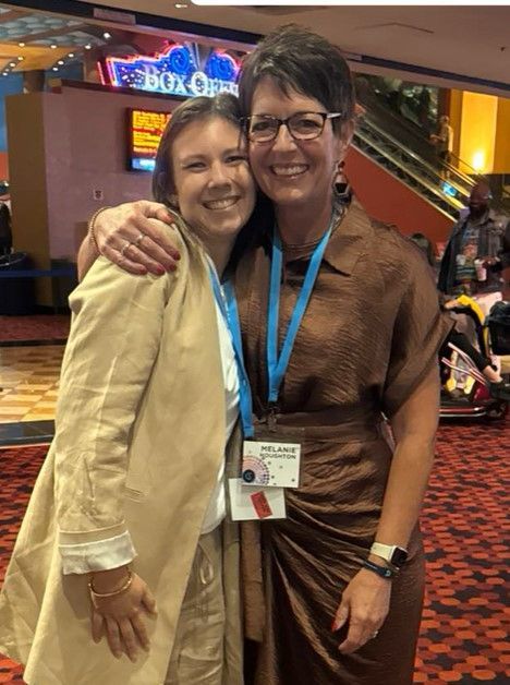 Two women are posing for a picture in front of a sign that says box office