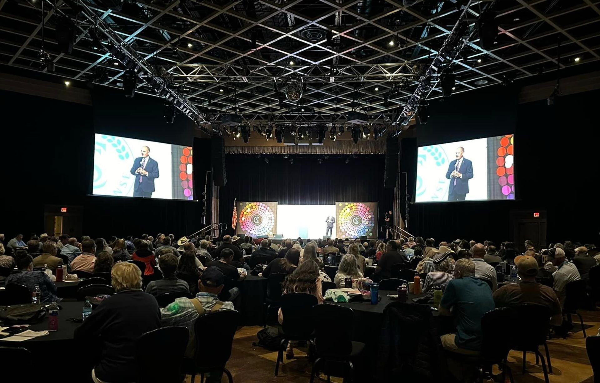 A large group of people are sitting at tables in front of a large screen.