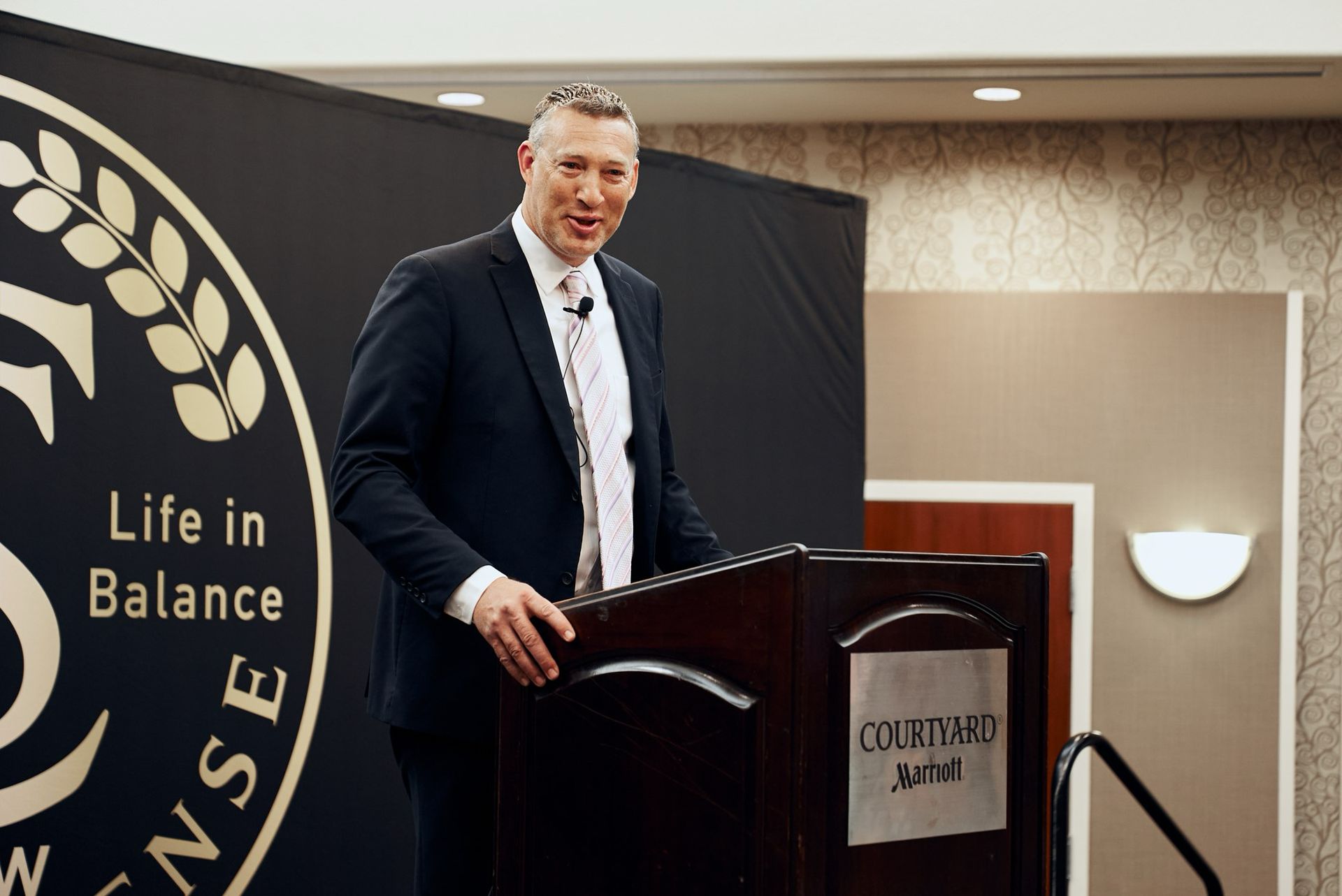 A man in a suit is standing at a podium in front of a sign that says life in balance
