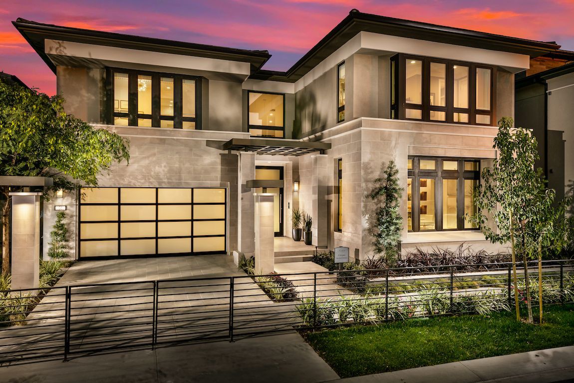 Modern two-story house with gray stucco facade, glass garage door, and lush landscaping against a sunset sky.