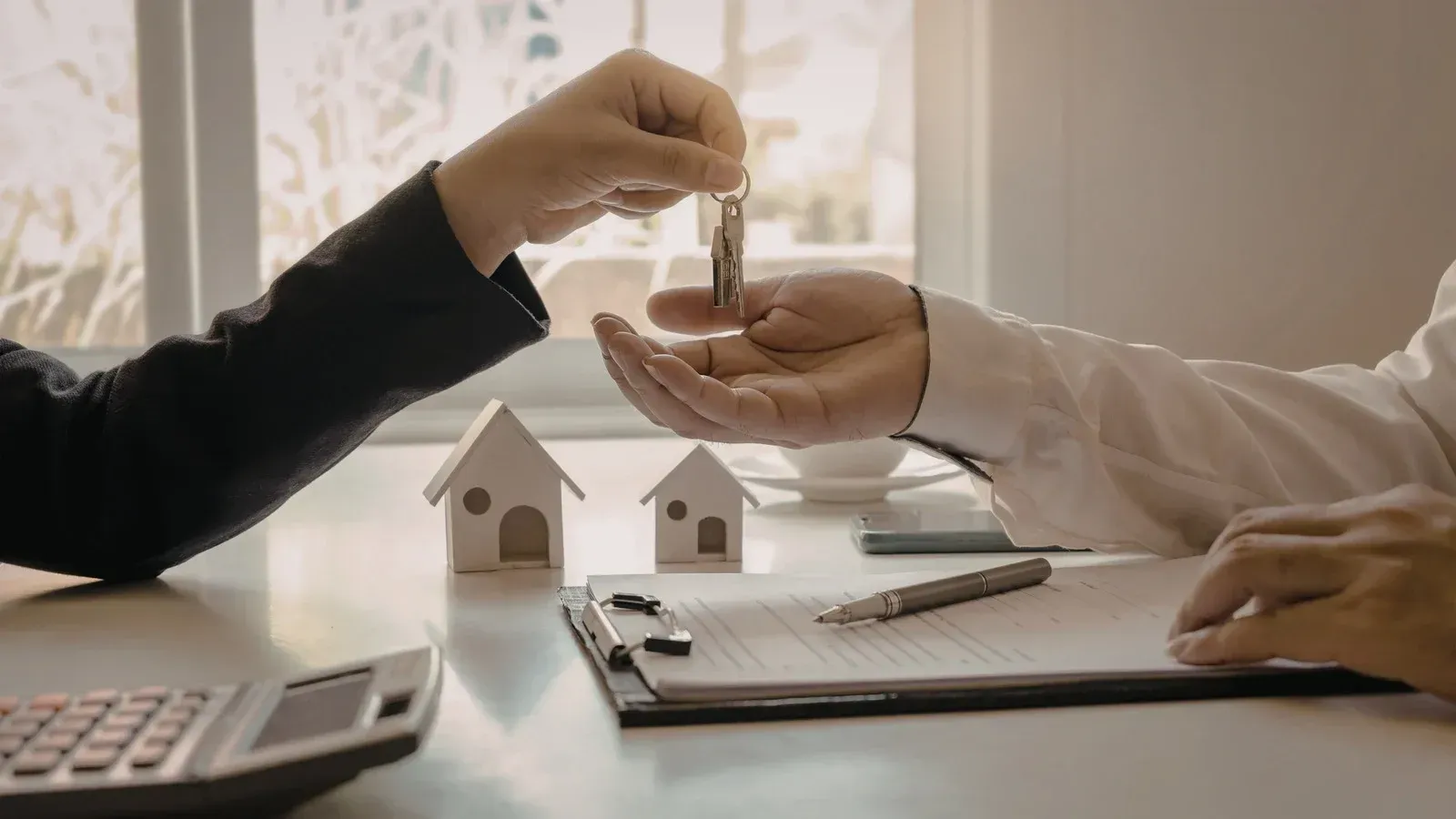 Person handing keys to another person over a desk with paperwork, toy houses, and a calculator.