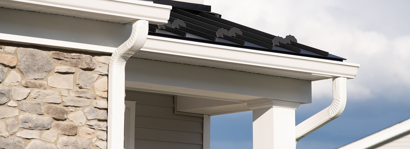 White gutter system on a building with stone siding and black roof. Blue sky is visible.
